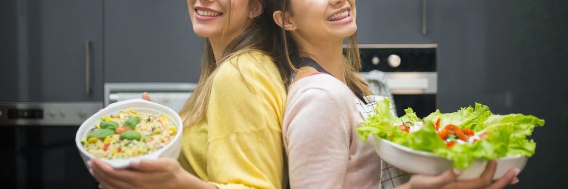 Two smiling women hold vibrant salads in a kitchen setting, showcasing fresh culinary creations.